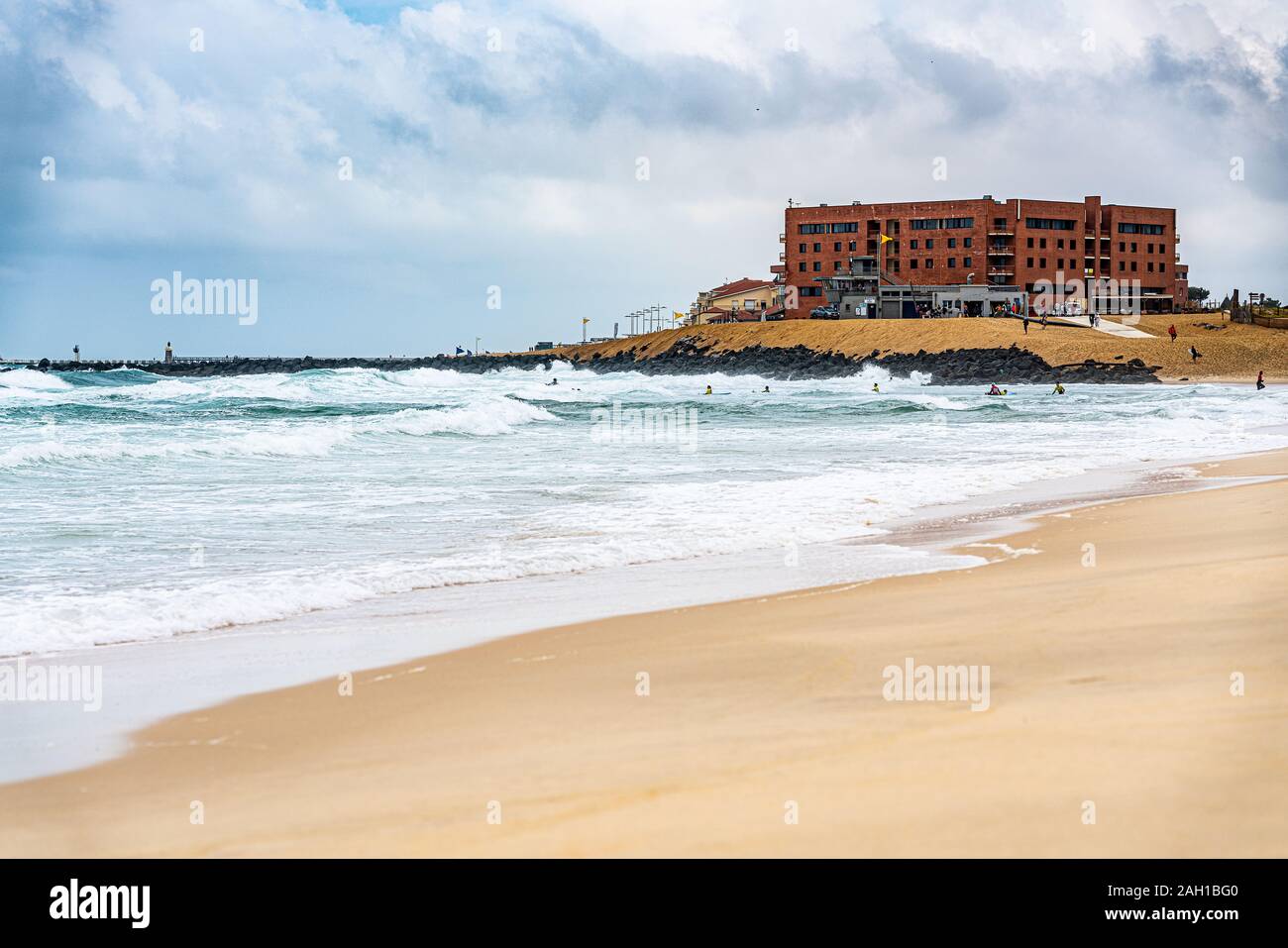 Bâtiment en briques rouges dans les dunes derrière la plage avec mer agitée et les vagues s'écraser dans les rochers, plage de vide sur le premier plan et de surfeurs à l'eau Banque D'Images