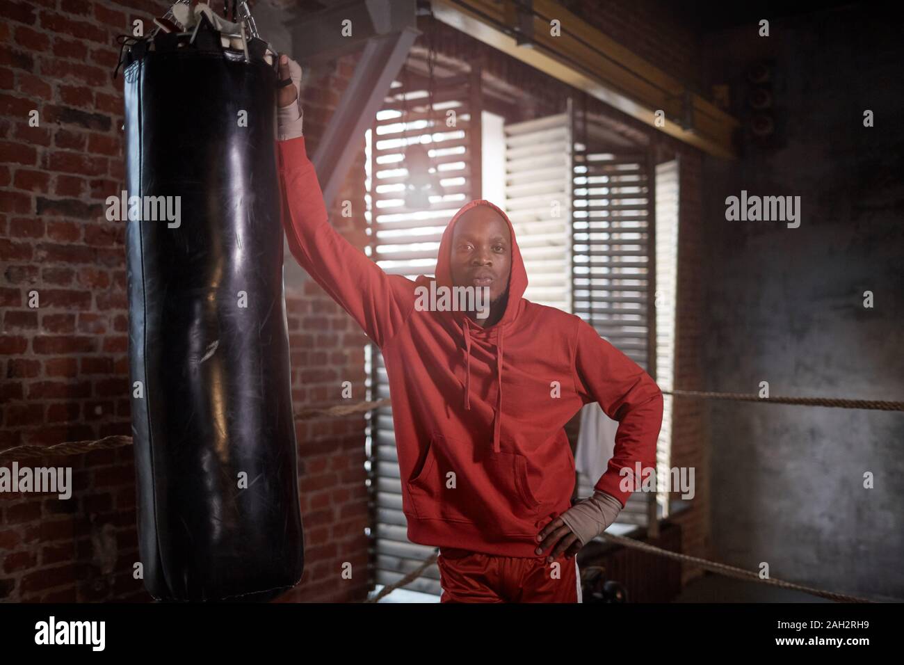 Portrait de jeune homme africain dans les vêtements de sport, debout près de l'entraînement de sac de boxe et looking at camera in gym Banque D'Images