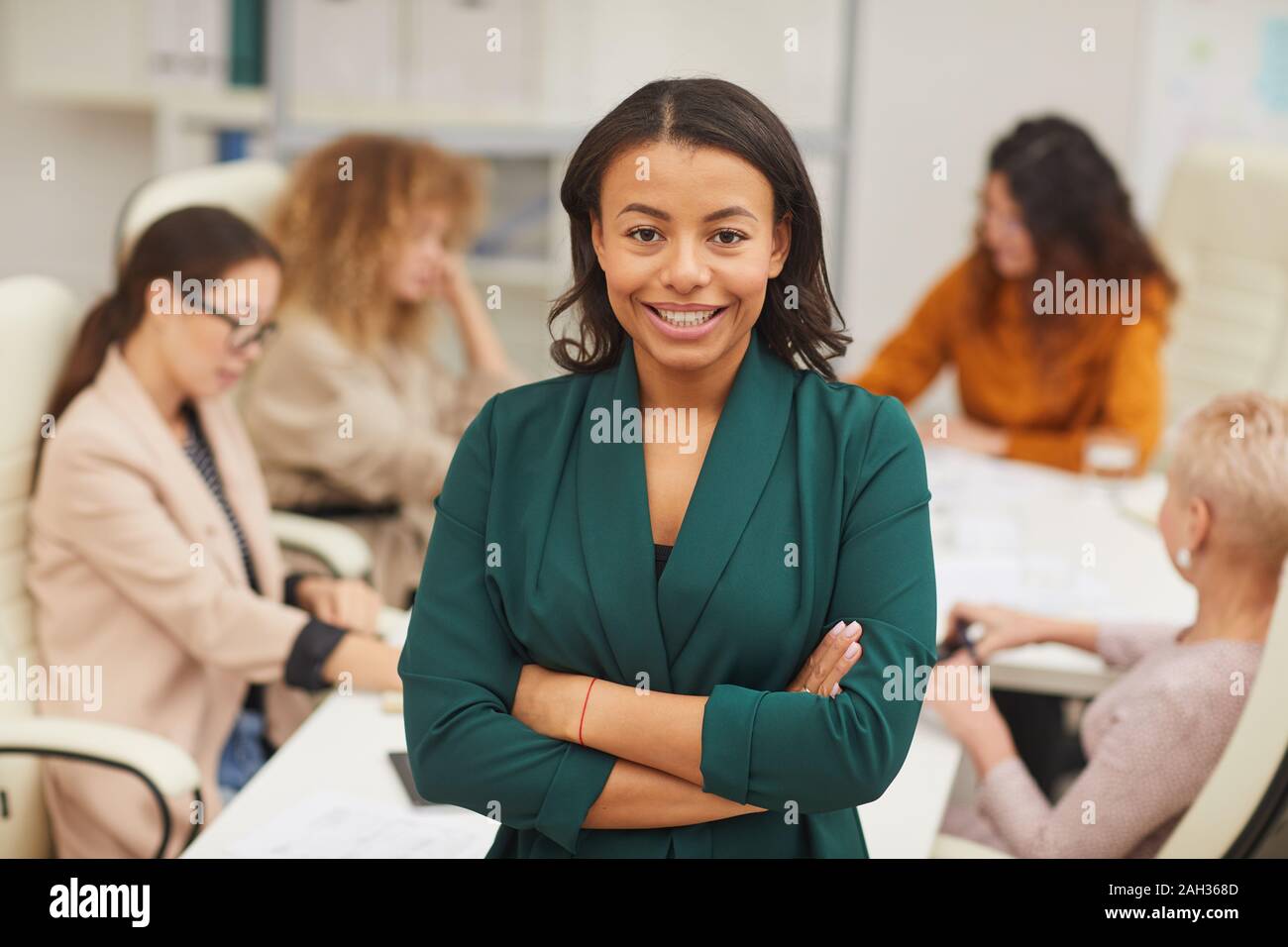 Charmant African American Woman standing with arms fermé à la caméra avec des collègues au sourire coworking derrière elle, jusqu'à la taille portrait Banque D'Images