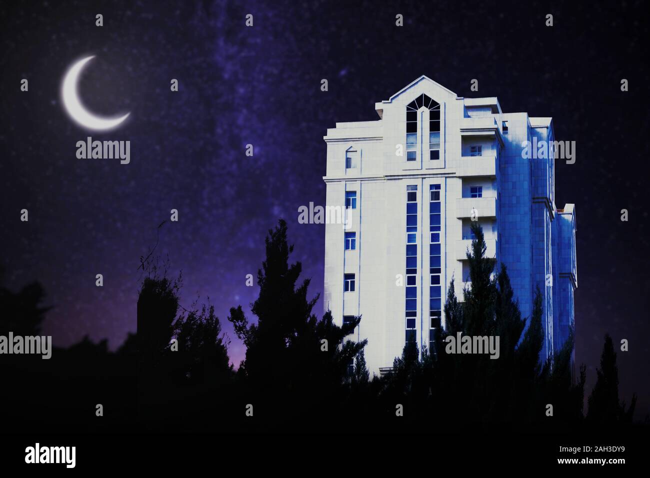 Une tour résidentielle blanc à Ashgabat, Turkménistan de nuit avec des silhouettes de sapins sur l'avant-plan et un croissant sur la nuit ciel stary. Banque D'Images