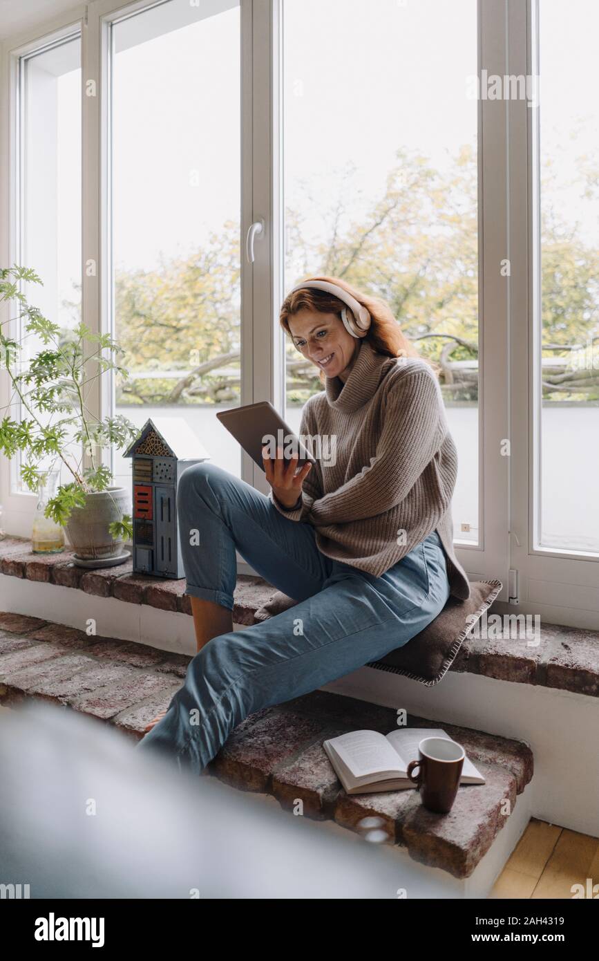 Mature Woman sitting on wondow sill, using digital tablet Banque D'Images