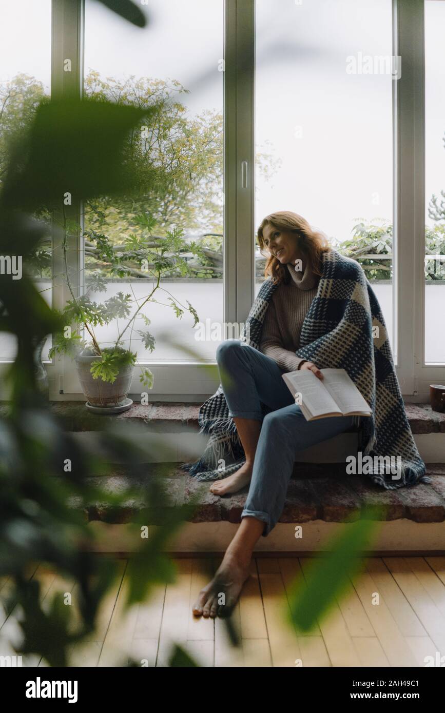 Mature Woman sitting on wondow sill, enveloppé dans une couverture, reading book Banque D'Images