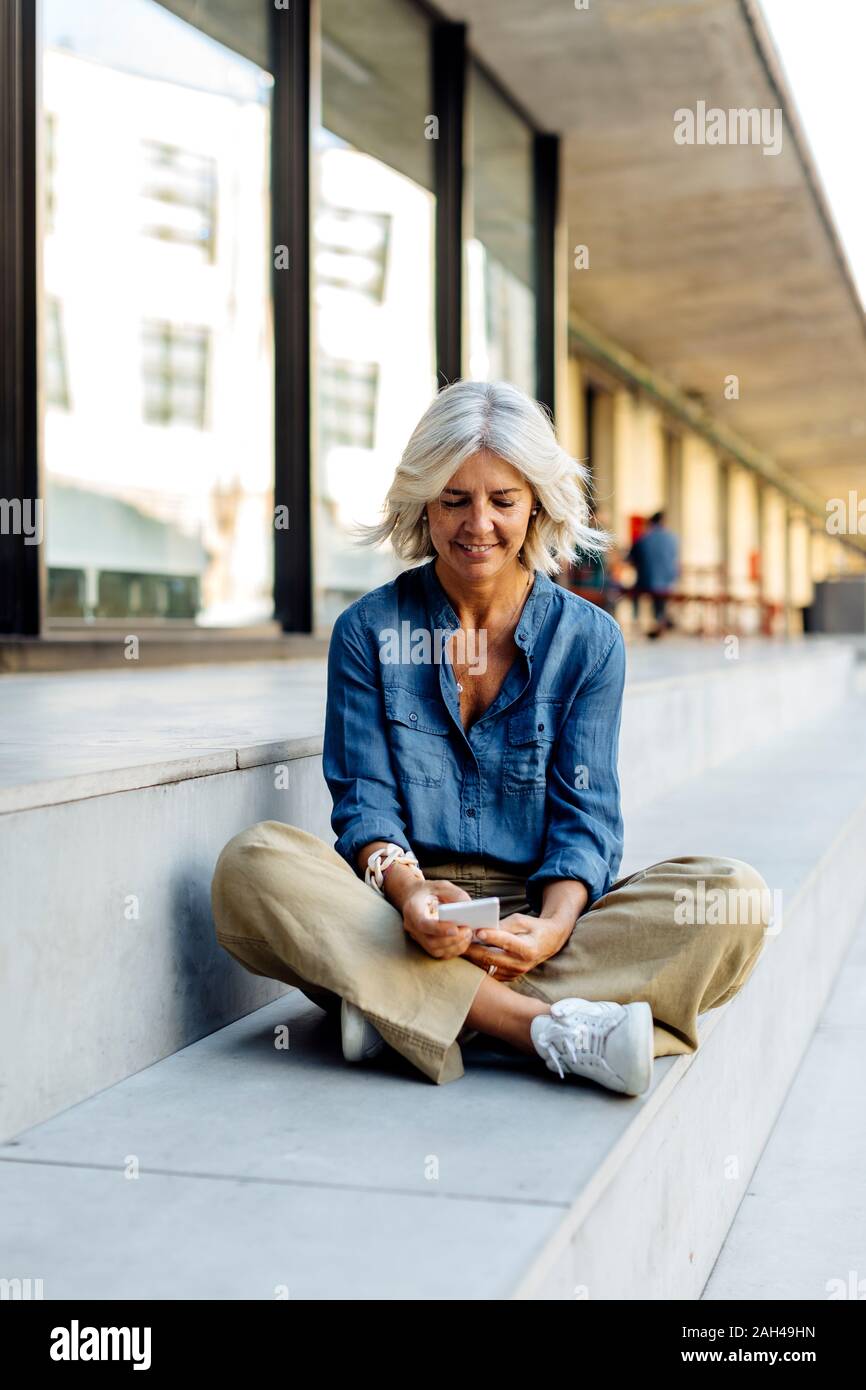 Femme d'âge mûr assis en tailleur sur des pas dans la ville, using smartphone Banque D'Images