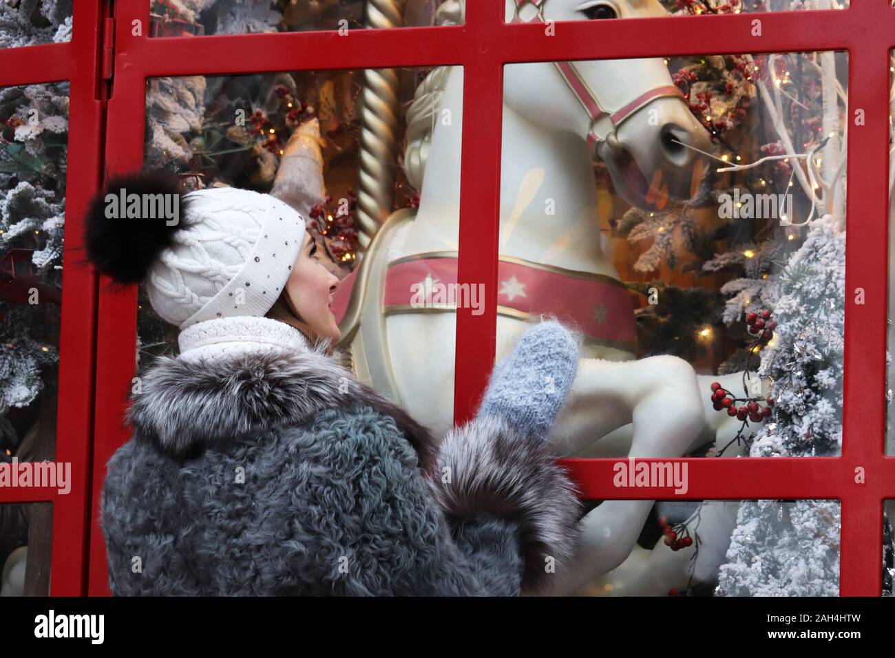 Happy girl in hat tricoté et mitaines à la boutique à la fenêtre avec cheval jouet et Nouvel An décorations. Magie de Noël Banque D'Images