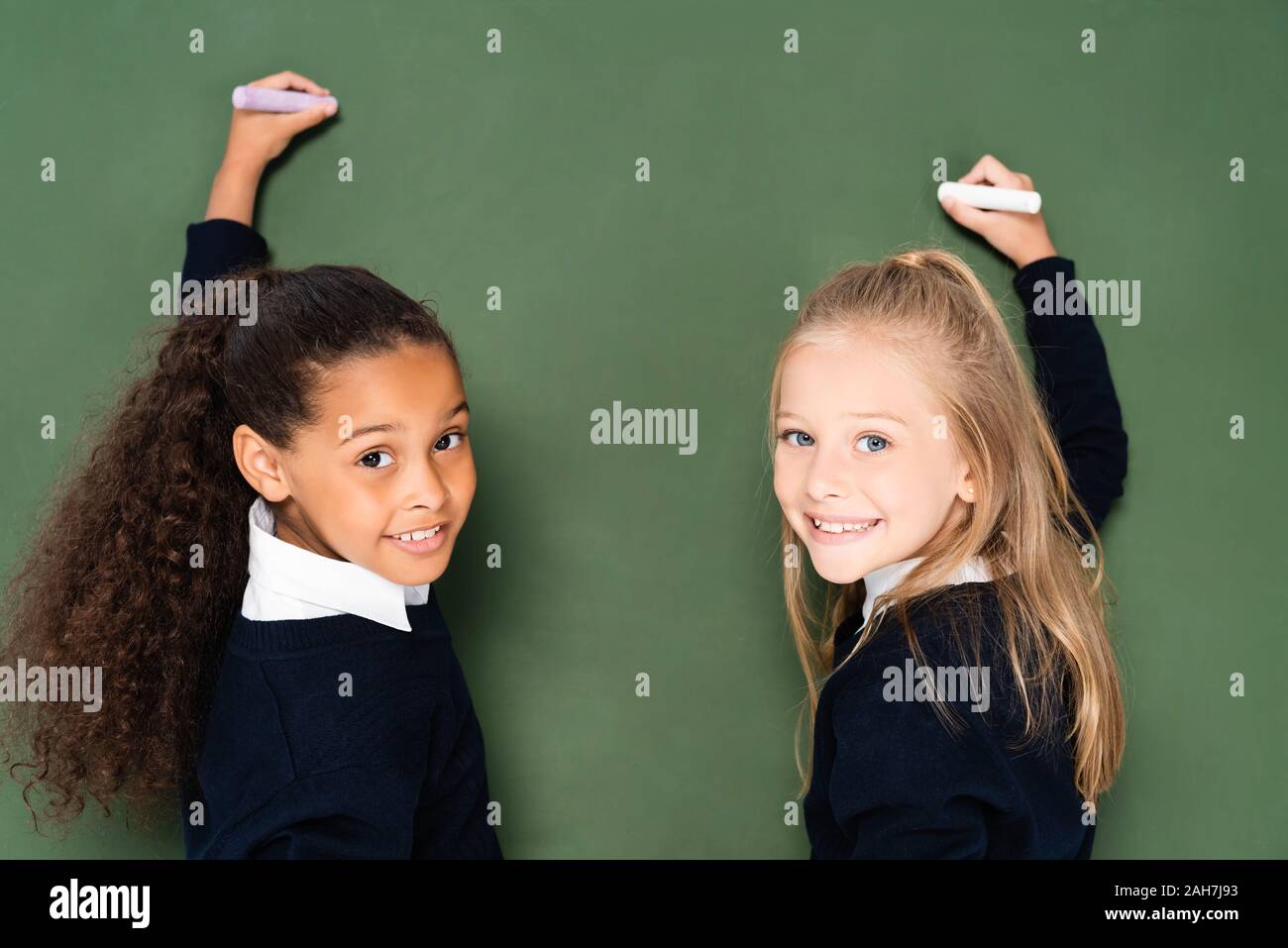 Deux écolières multiculturelle joyeuse smiling at camera while writing on chalkboard Banque D'Images