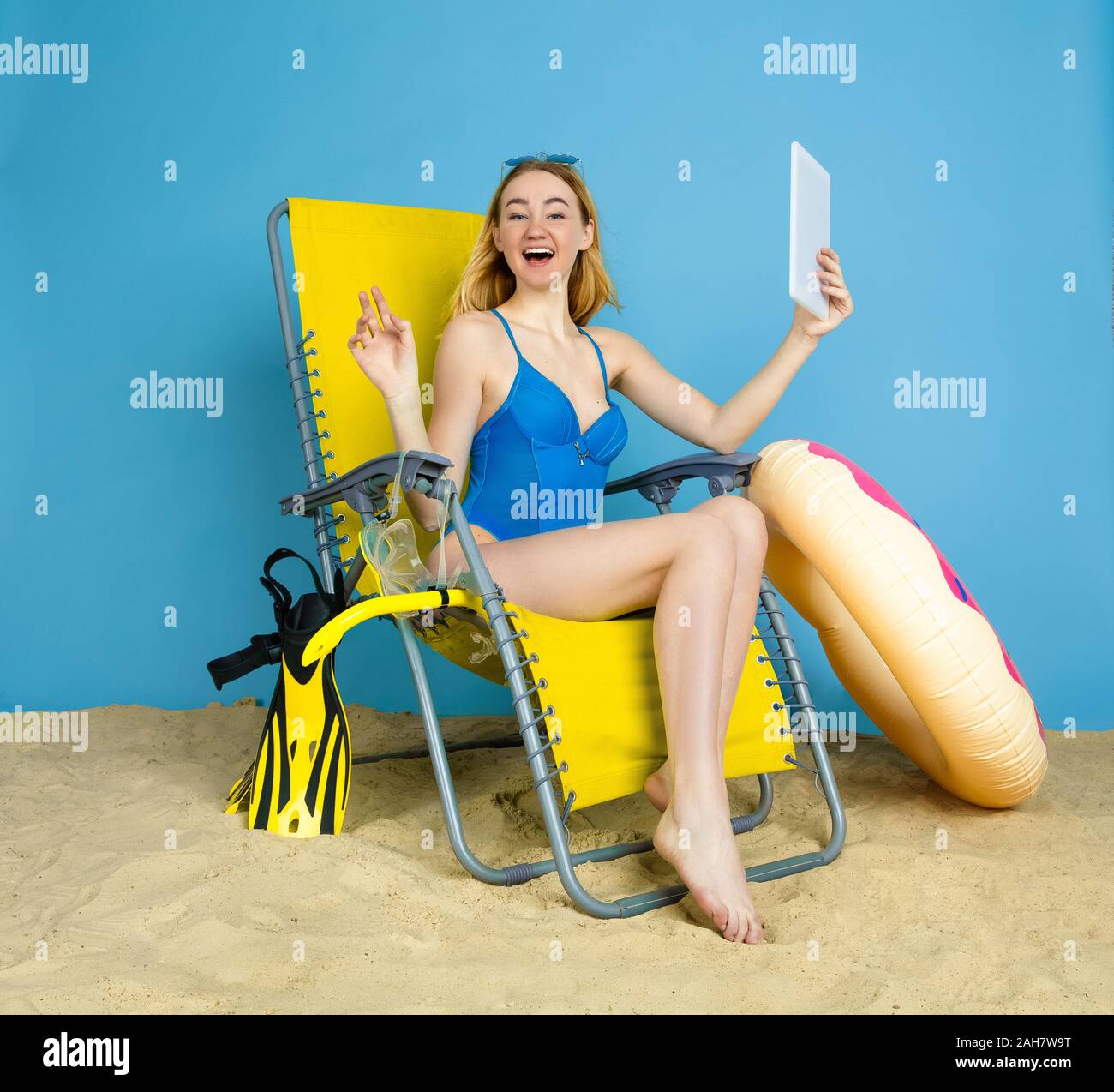 Happy young woman with tablet selfies prend ou vlog sur les voyages sur fond studio bleu. Concept d'émotions humaines, l'expression du visage, vacances, week-end d'été. L'été, mer, océan, l'alcool. Banque D'Images