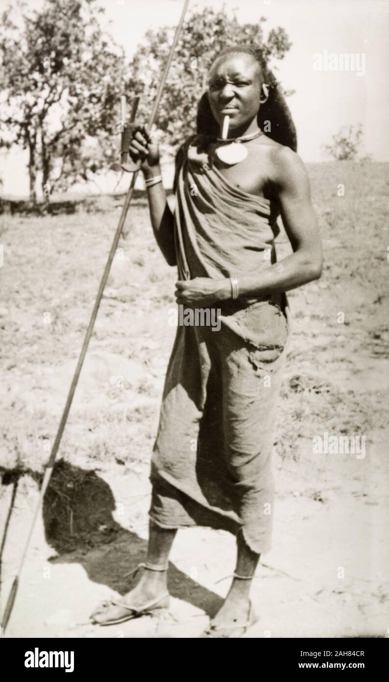Kenya, portrait en pied d'un homme en costume traditionnel, orné de bijoux et posant avec une lance, 1933-1934. 1995/076/1/1/23/12. Banque D'Images