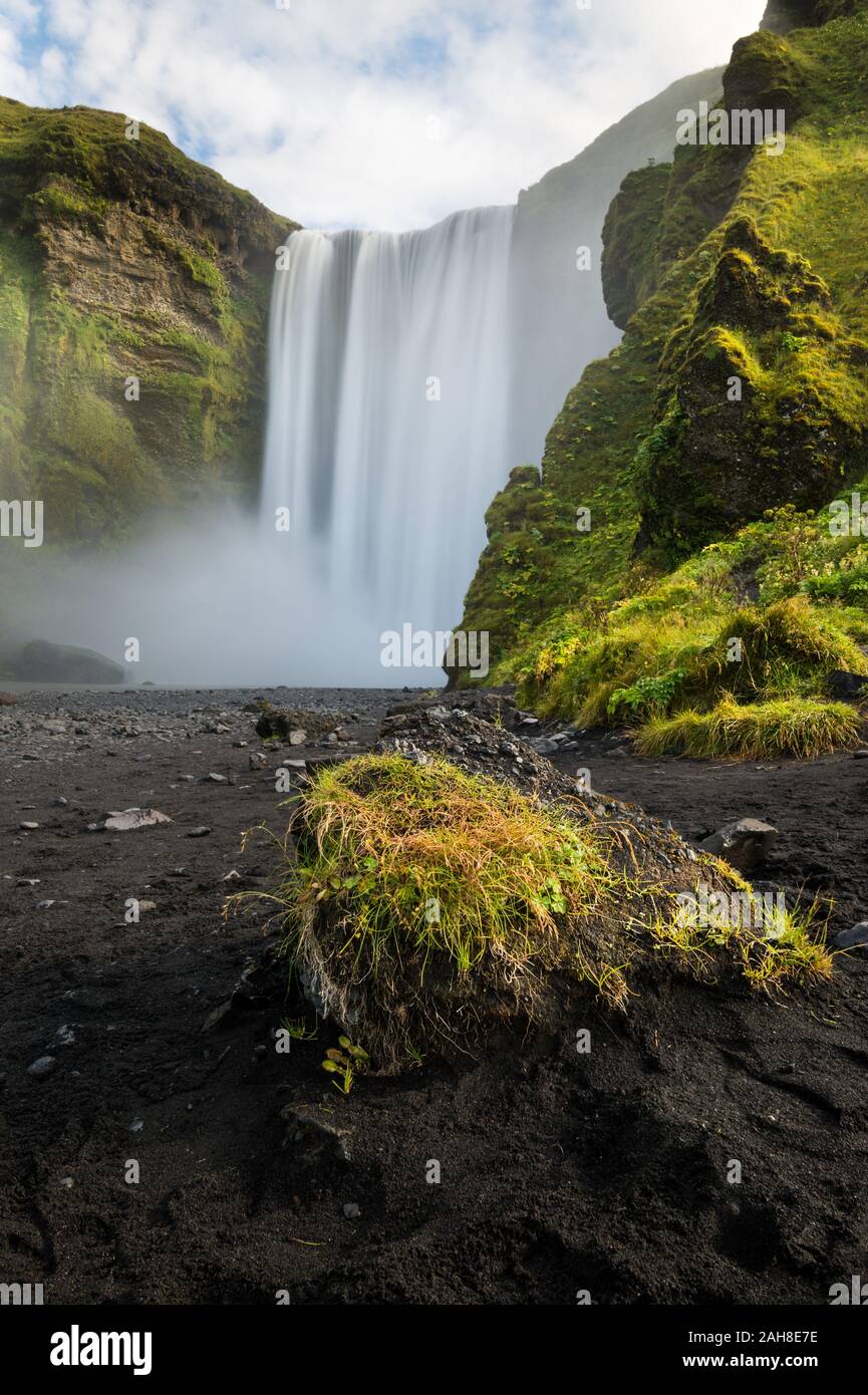 Vue à bas angle de la cascade géante de Skogafoss, qui coule entre la roche recouverte de mousse et une plage de sable noir Banque D'Images