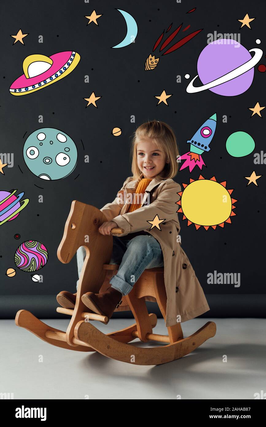 Heureux l'enfant en trench coat et jeans assis sur cheval à bascule sur fond noir avec illustration cosmique fée Banque D'Images