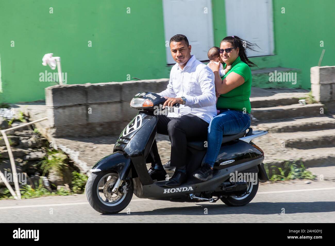 L'image spectaculaire de deux adultes et un enfant à cheval sur un scooter dans un centre-ville ville de montagne des Caraïbes. Banque D'Images