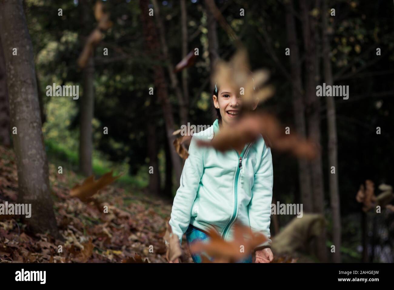 Petite fille souriante regardant des feuilles brunes voler dans la forêt. Gamin ludique avec veste polaire verte s'amuser dans les bois le jour de l'automne Banque D'Images