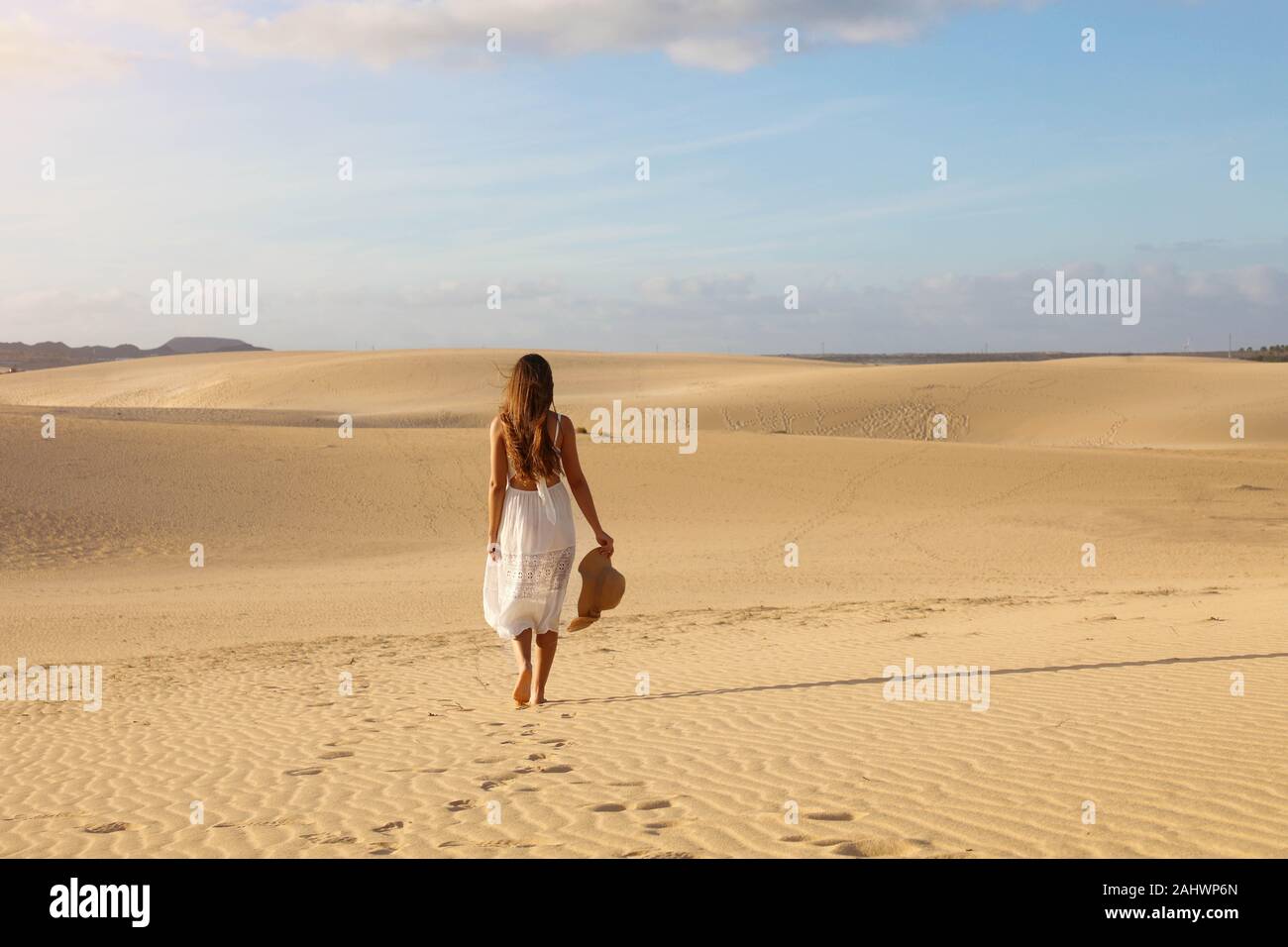 Belle jeune femme avec robe blanche marche dans les dunes du désert au coucher du soleil. Fille qui marche sur le sable doré sur Corralejo Beach, à Fuerteventura. Banque D'Images