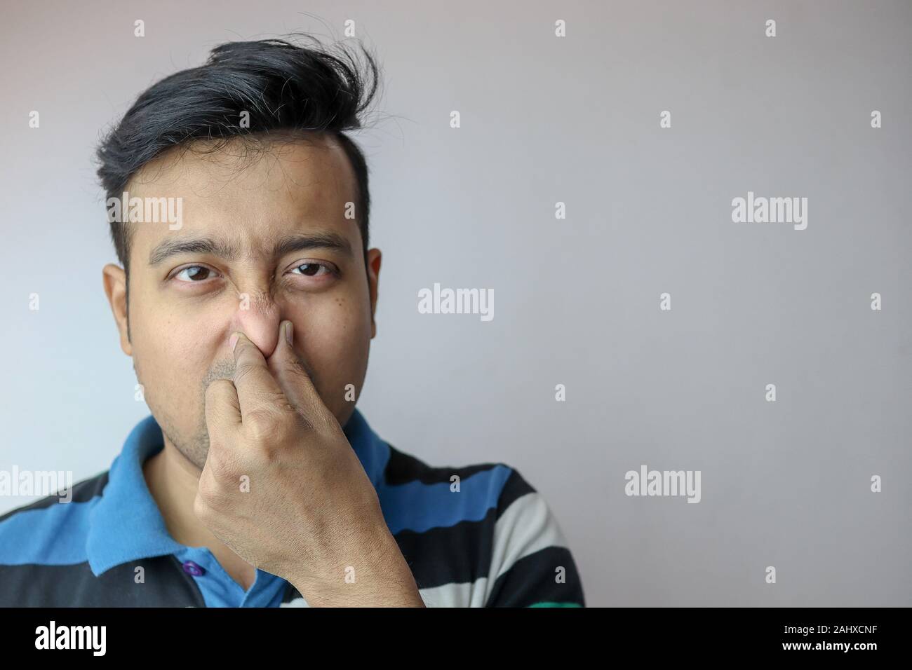 Un jeune indien homme montrant le geste de l'odeur fétide de pincer le nez avec les mains et les sourcils isolé sur gris avec copie espace pour le texte Banque D'Images