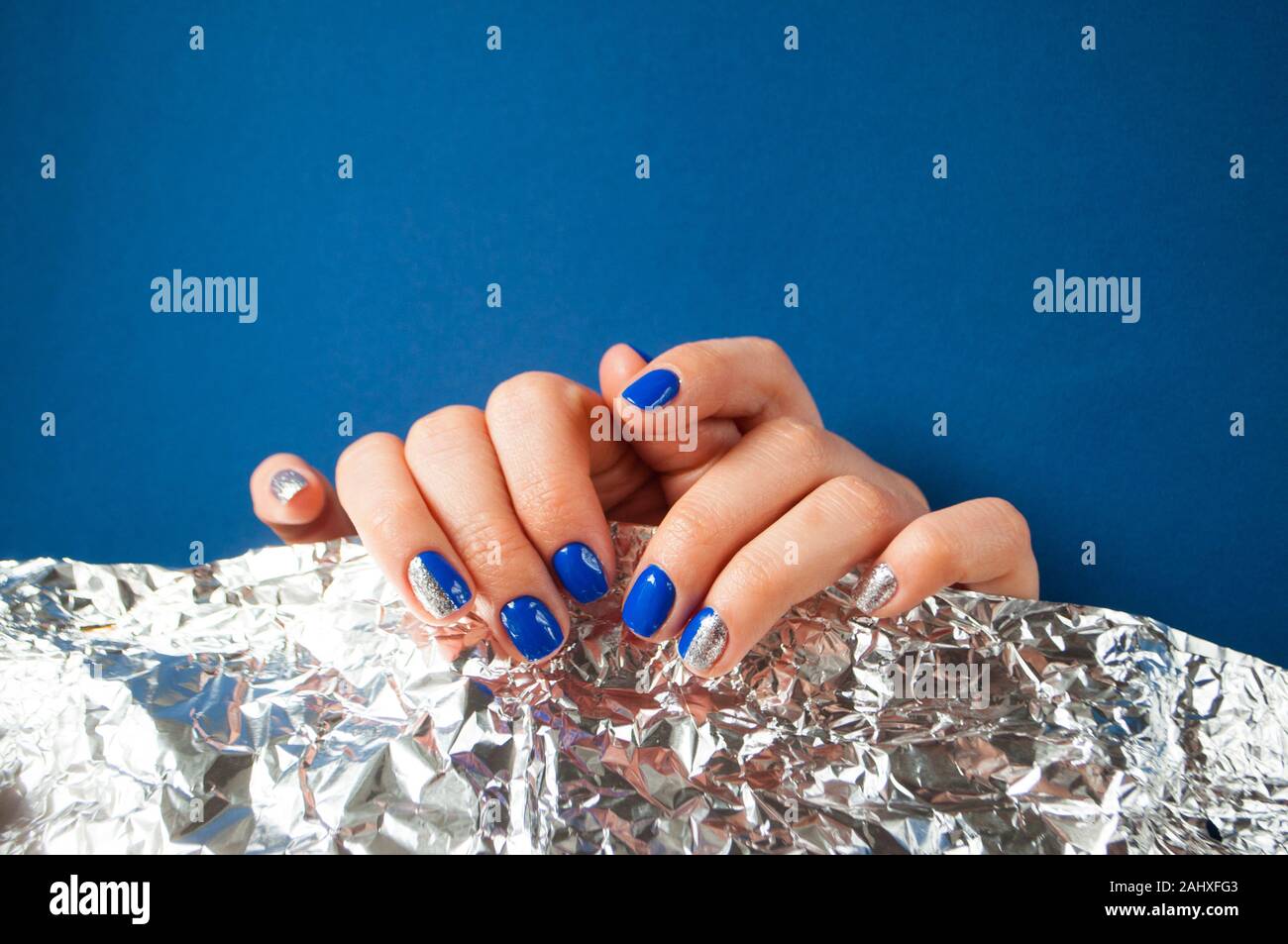 Jeune femme ses mains avec de beaux ongles bleu sur fond bleu avec du papier brillant métallique. Couleur 2020 tendance. Banque D'Images
