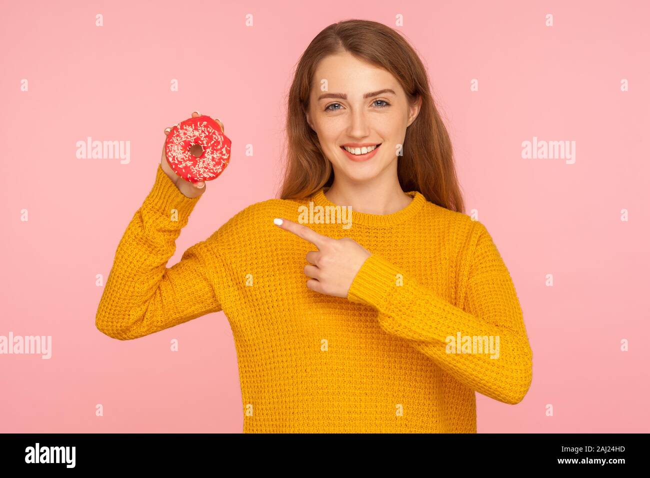 Regardez donut ! Portrait of smiling cute girl à gingembre sweater pointant vers donut rose and smiling at camera, montrant, de confiserie de sucre sucré j Banque D'Images