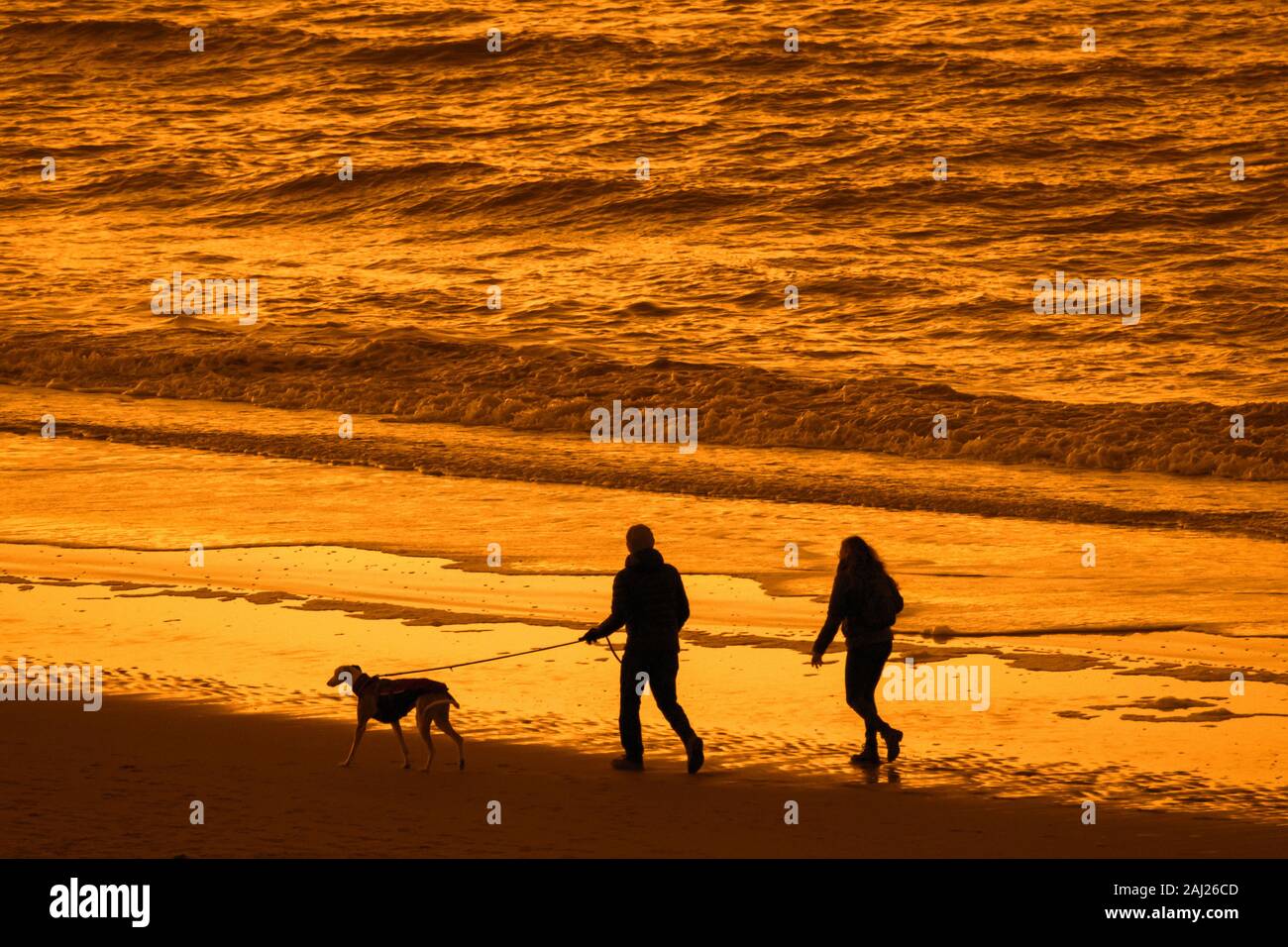 Couple de propriétaires de chien à marcher le long de la côte de la mer du Nord avec whippet chien sur une plage de sable, en silhouette au coucher du soleil sur une soirée froide en hiver Banque D'Images