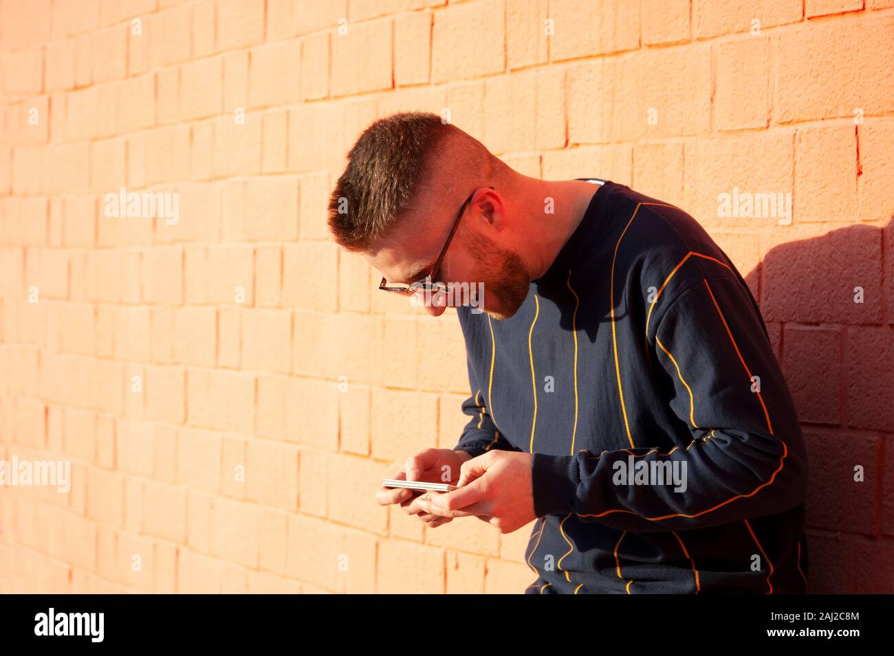 Guy smiling positive dans casualwear avec téléphone mobile. Young caucasian man standing at outdoor mur rouge et voir des vidéos drôles dans son appareil. Loisirs, fu Banque D'Images