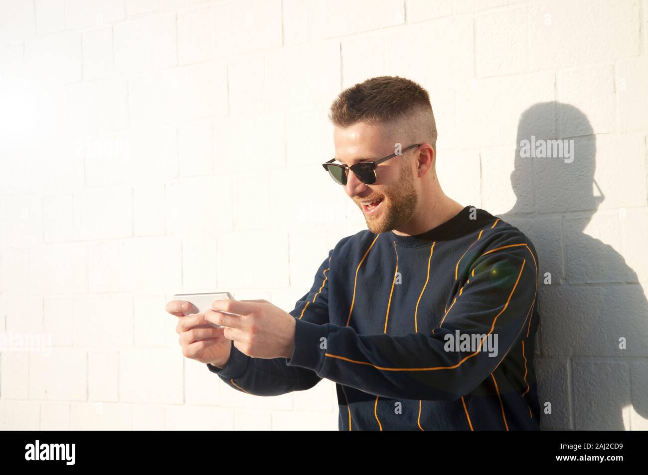 Guy smiling positive dans casualwear avec téléphone mobile. Young caucasian man standing at outdoor mur blanc et voir des vidéos drôles dans son appareil. Les loisirs, Banque D'Images