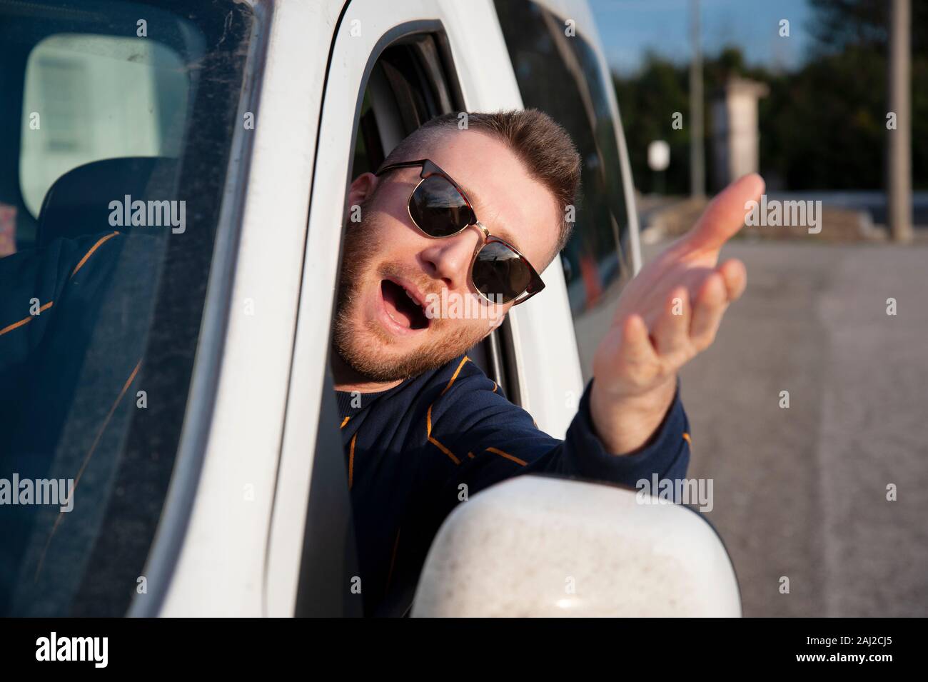 Jeune homme avec des lunettes qui sort de la fenêtre de voiture avec sa tête  avec son bras levé. Expression de visage en colère et disputes de pilote  des gestes. Test EXPRES