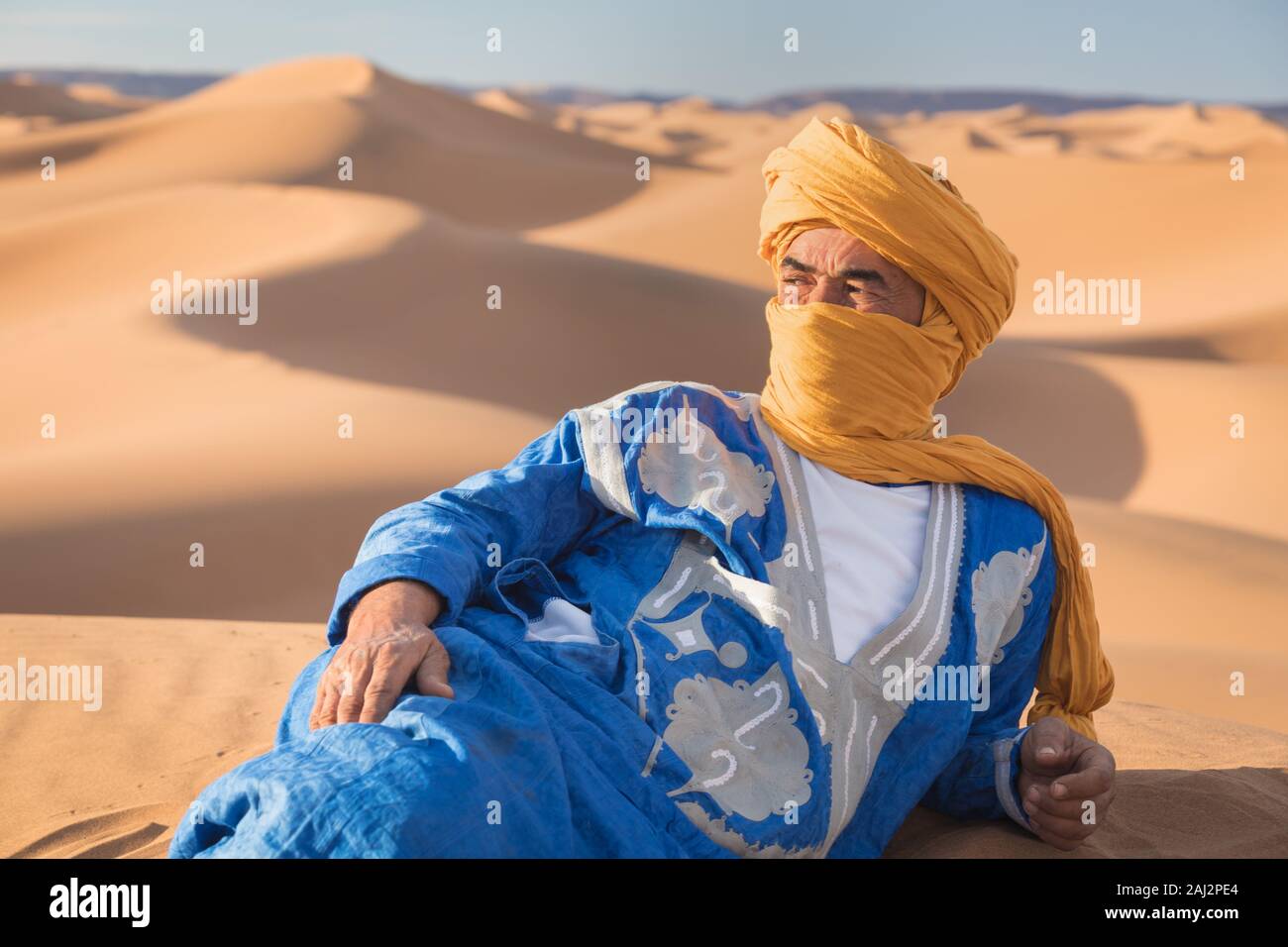 Portrait d'un Bédouin nomad avec turban coloré et grand sourire assis sur dune de sable touristique populaire Merzouga désert. Homme Touareg Banque D'Images