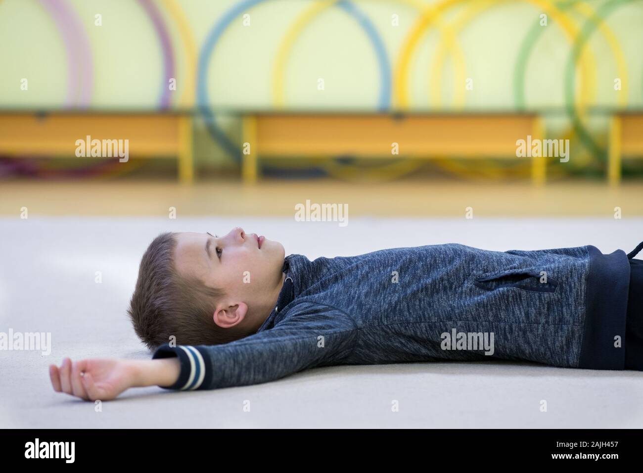 Jeune enfant garçon fixant et relaxiong alors qu'elle repose sur le plancher à l'intérieur de la salle de sport dans une école après la formation. Banque D'Images