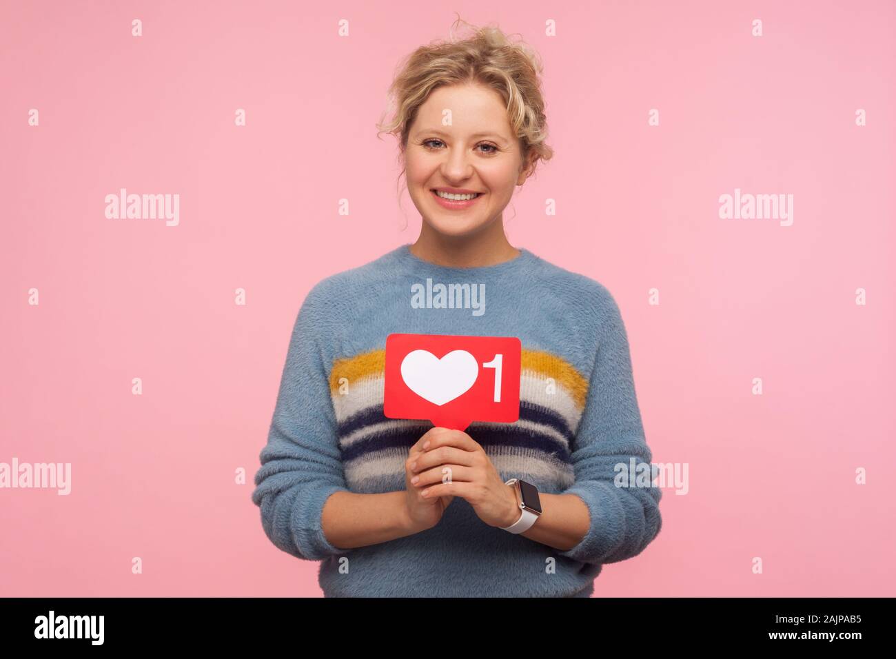 Portrait of happy young woman avec les cheveux bouclés pull-over chaud holding heart comme icon, aiment le contenu, symbole de notification de galet, un bouton pour la m Banque D'Images