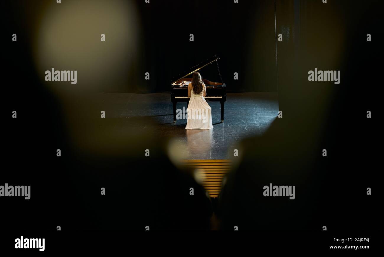 Une jeune fille vêtue de blanc avec son dos à l'auditoire joue un grand piano noir sur une scène. Banque D'Images