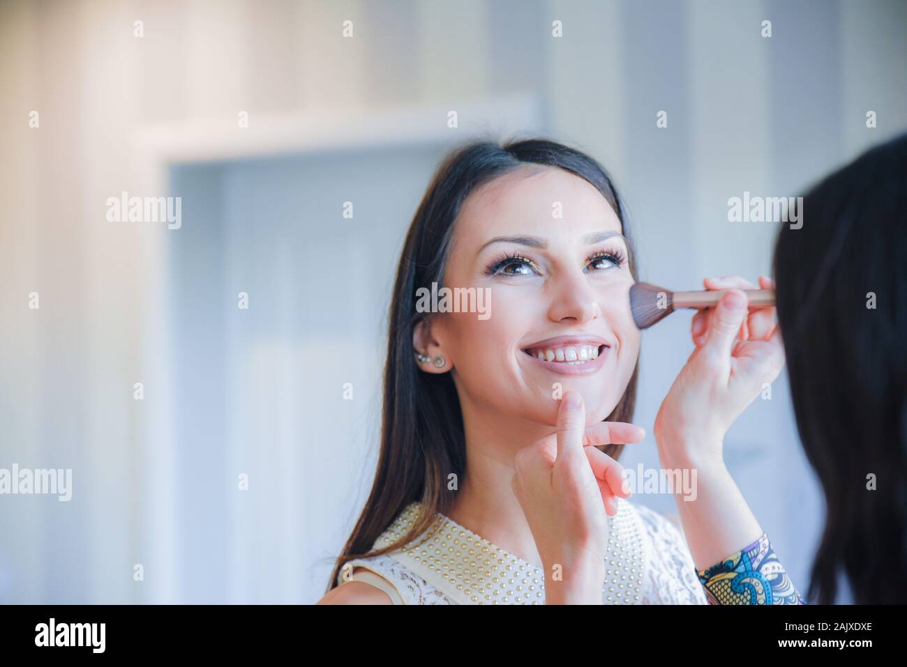 Belle jeune fille brunette souriante et esthétique tout en obtenant son visage tonicité avec un pinceau doux par un artiste professionnel de maquillage Banque D'Images