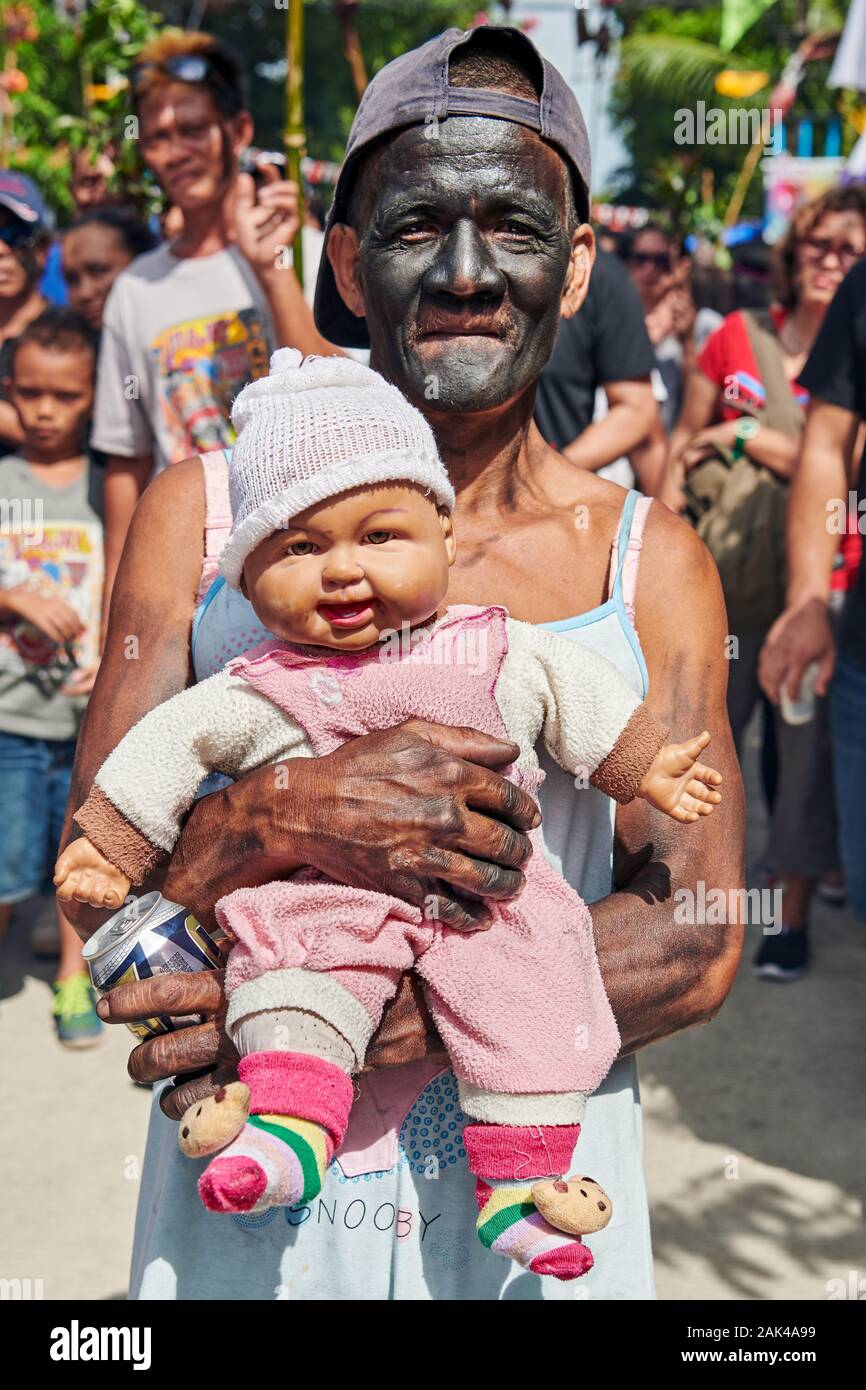 Ville d'Ibajay, province d'Aklan, Philippines: Vieil homme à visage noir tenant une poupée de bébé à la parade de rue Du festival Ati-Atihan Banque D'Images