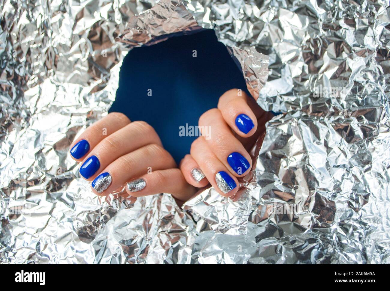 Jeune femme ses mains avec de beaux ongles bleu sur fond bleu avec du papier brillant métallique. Couleur 2020 tendance. Banque D'Images