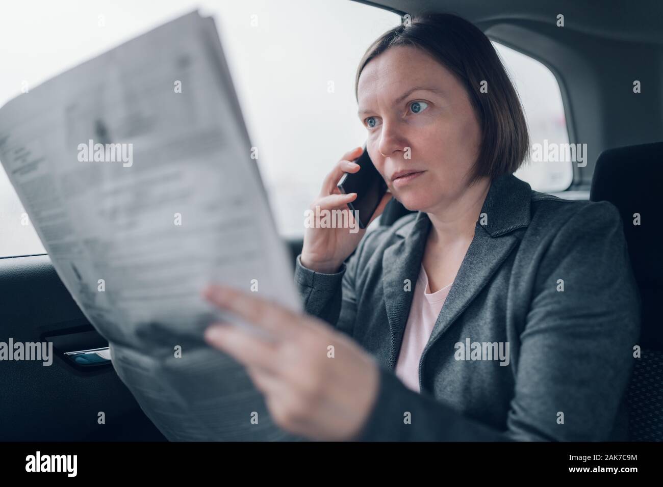 Businesswoman reading newspaper et à l'aide de téléphone mobile à l'arrière de voiture lors des trajets au travail, selective focus Banque D'Images
