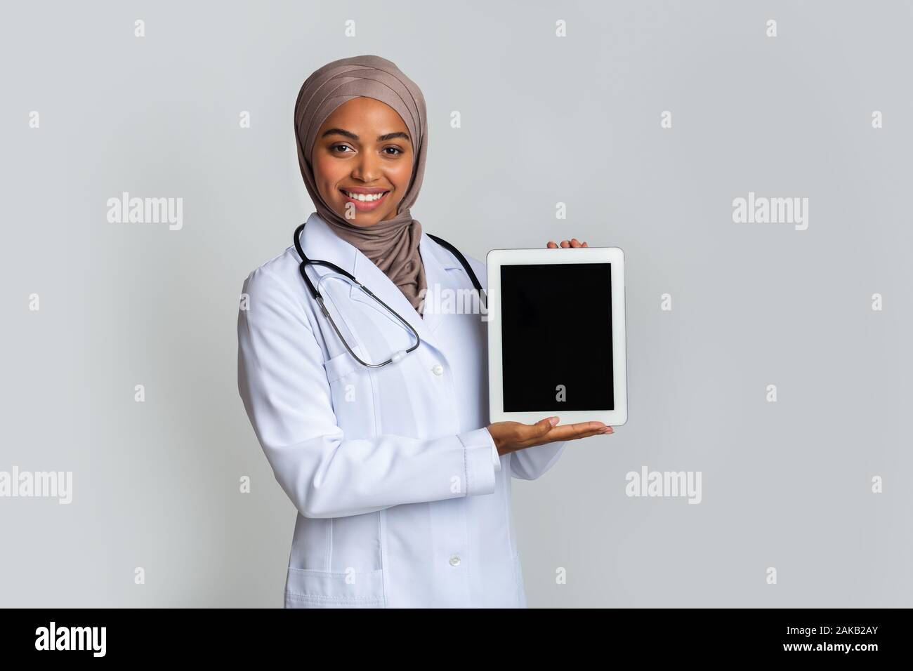 Pharmacie en ligne concept. Femme médecin musulmane noire holding digital tablet noir avec écran vide pour immersive, posant sur fond gris, l'espace libre Banque D'Images