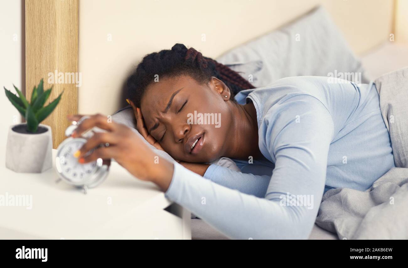 Un sommeil de mauvaise qualité. Alarm-Clock Afro femme tournant la tête hors de toucher le Réveil au lit dans la chambre. Selective Focus Banque D'Images