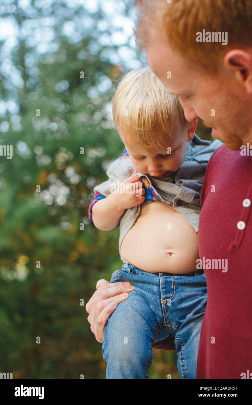 Un petit garçon dans les bras de papa soulève sa chemise pour vérifier son estomac Banque D'Images