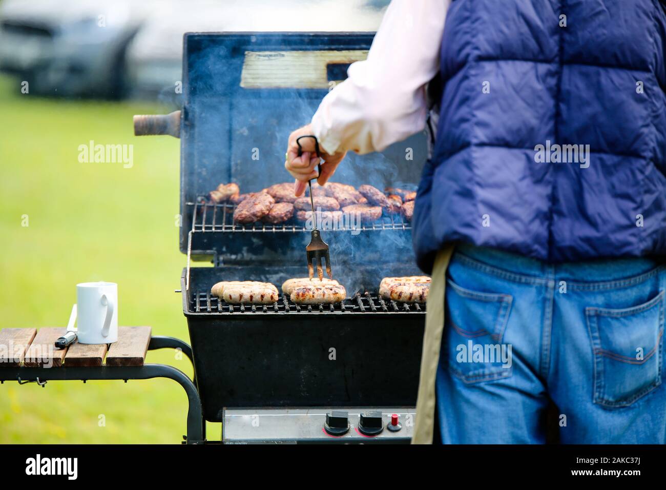 Un homme la cuisson des saucisses sur un barbecue à gaz ouvert, dans l'arrière-plan il y a un tas de hamburgers cuits en attente d'être servis aux clients Banque D'Images