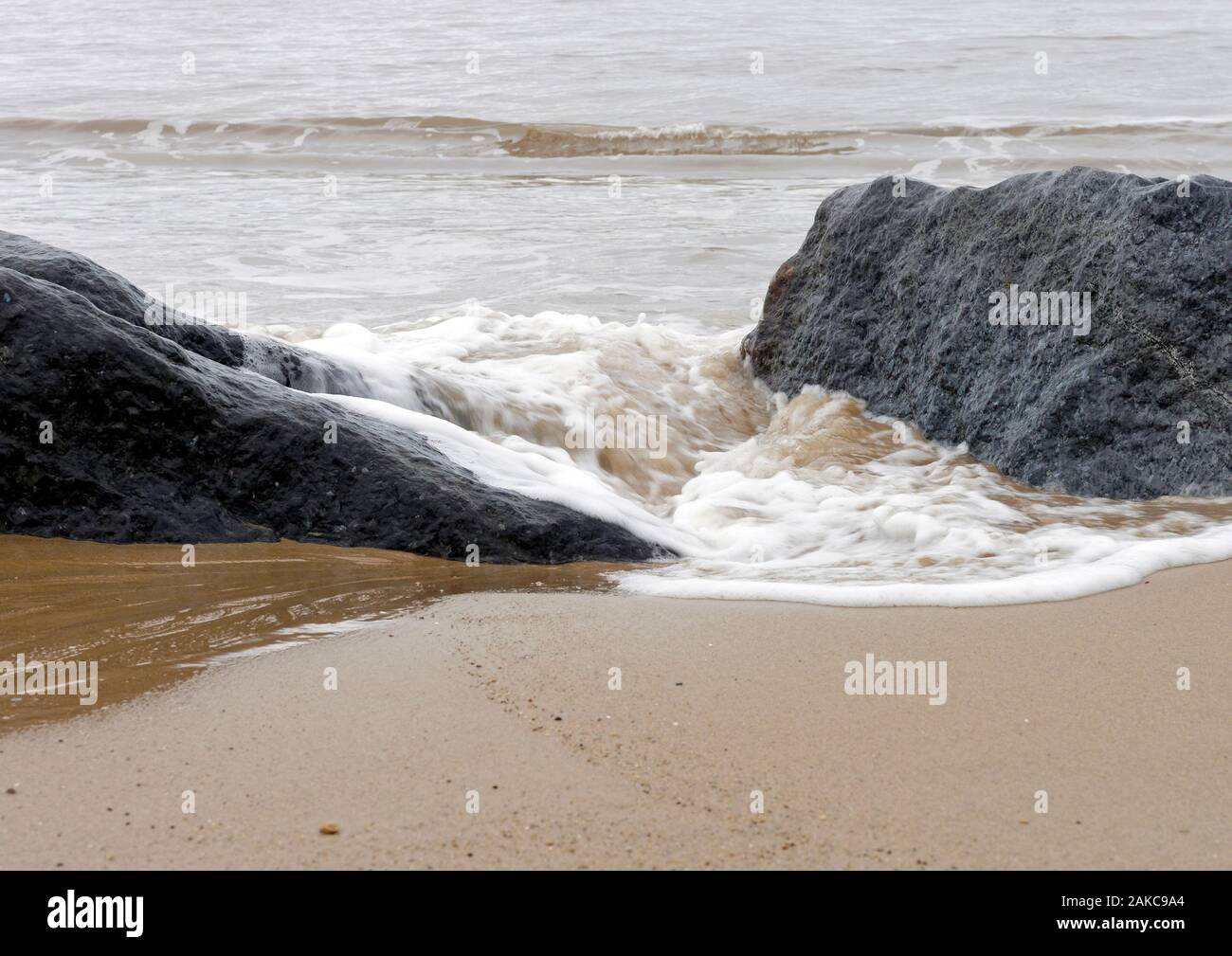 Sable, mer et rock - une douce vague éclabousse autour de grosses pierres placées sur une plage de Norfolk dans le cadre de la défense de la mer. Banque D'Images