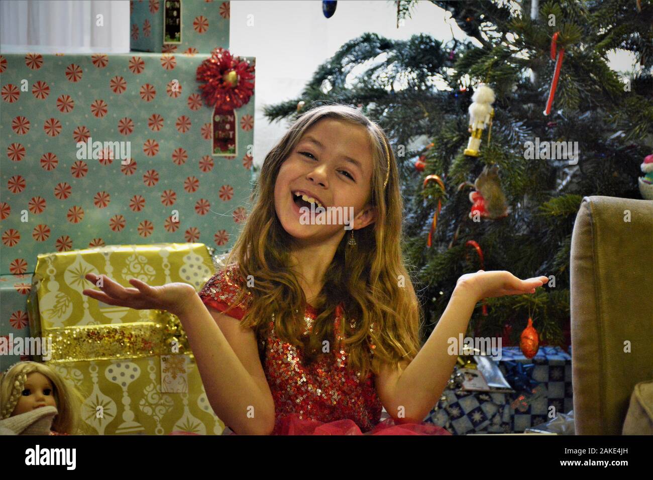 Fille de 7 ans à l'heure de Noël avec des cadeaux réels, certains pour elle, certains avec un arc dans ses cheveux et un arbre en arrière-plan Banque D'Images
