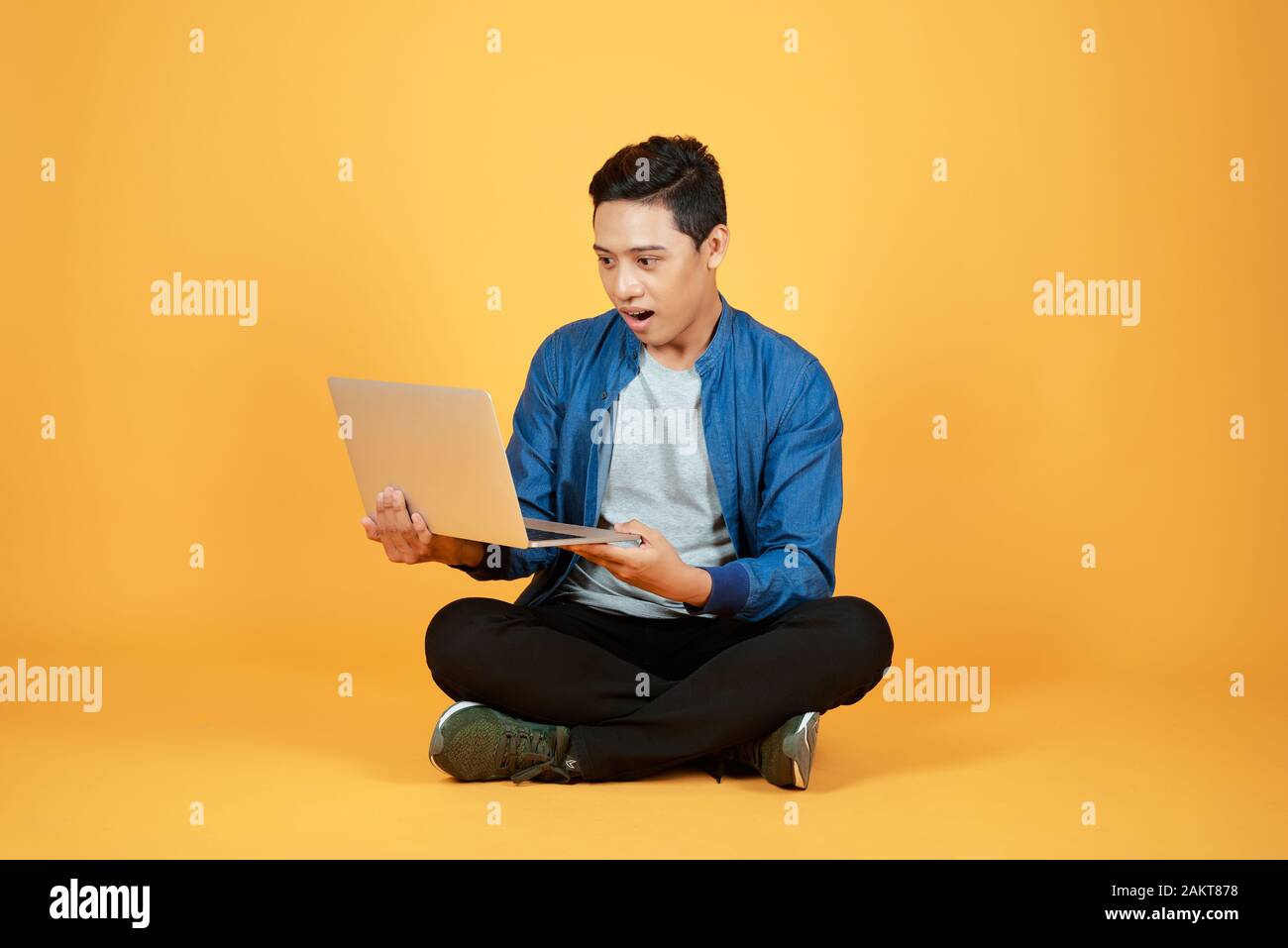 Jeune homme assis sur le sol et de travailler avec un ordinateur portable isolé sur fond orange en couleur. Banque D'Images