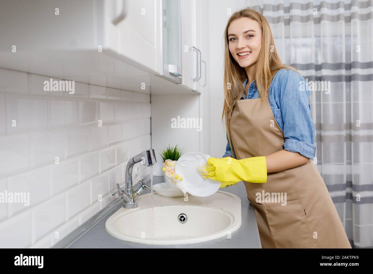Jolie fille blonde dans un tablier et gants jaunes laver la vaisselle dans une cuisine légère Banque D'Images