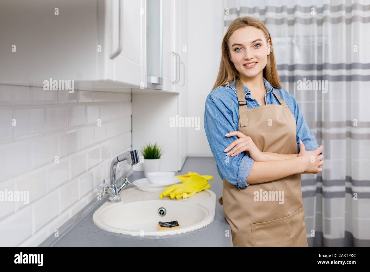 Blonde jeune femme souriante en tablier posant dans la cuisine, les plats sont lavés Banque D'Images
