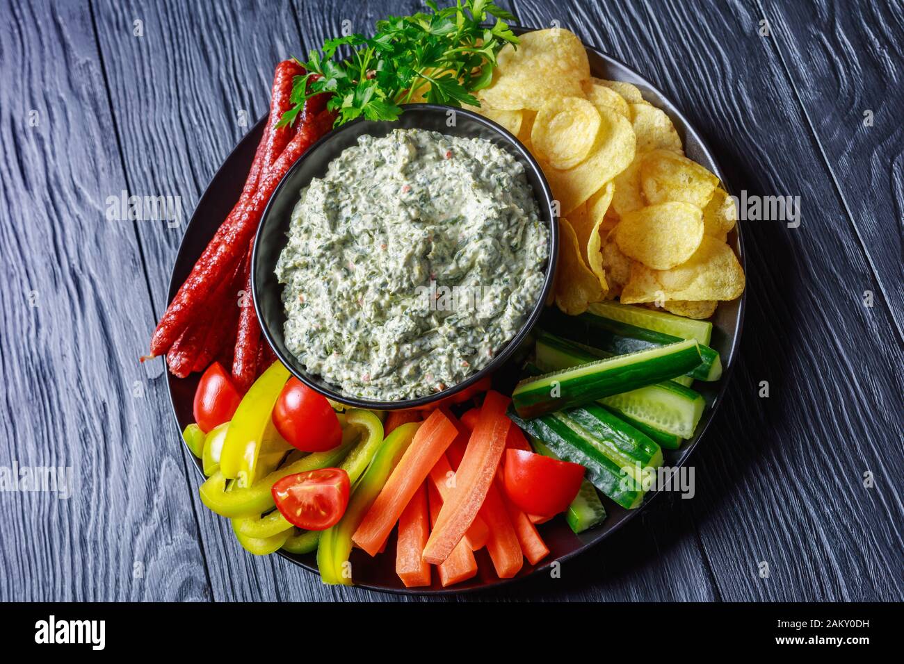 Les épinards crémeux trempent dans un bol avec saucisses, carottes, concombre, bâtonnets de poivron, chips de pommes de terre et persil frais sur une assiette, vue de dessus, planla Banque D'Images