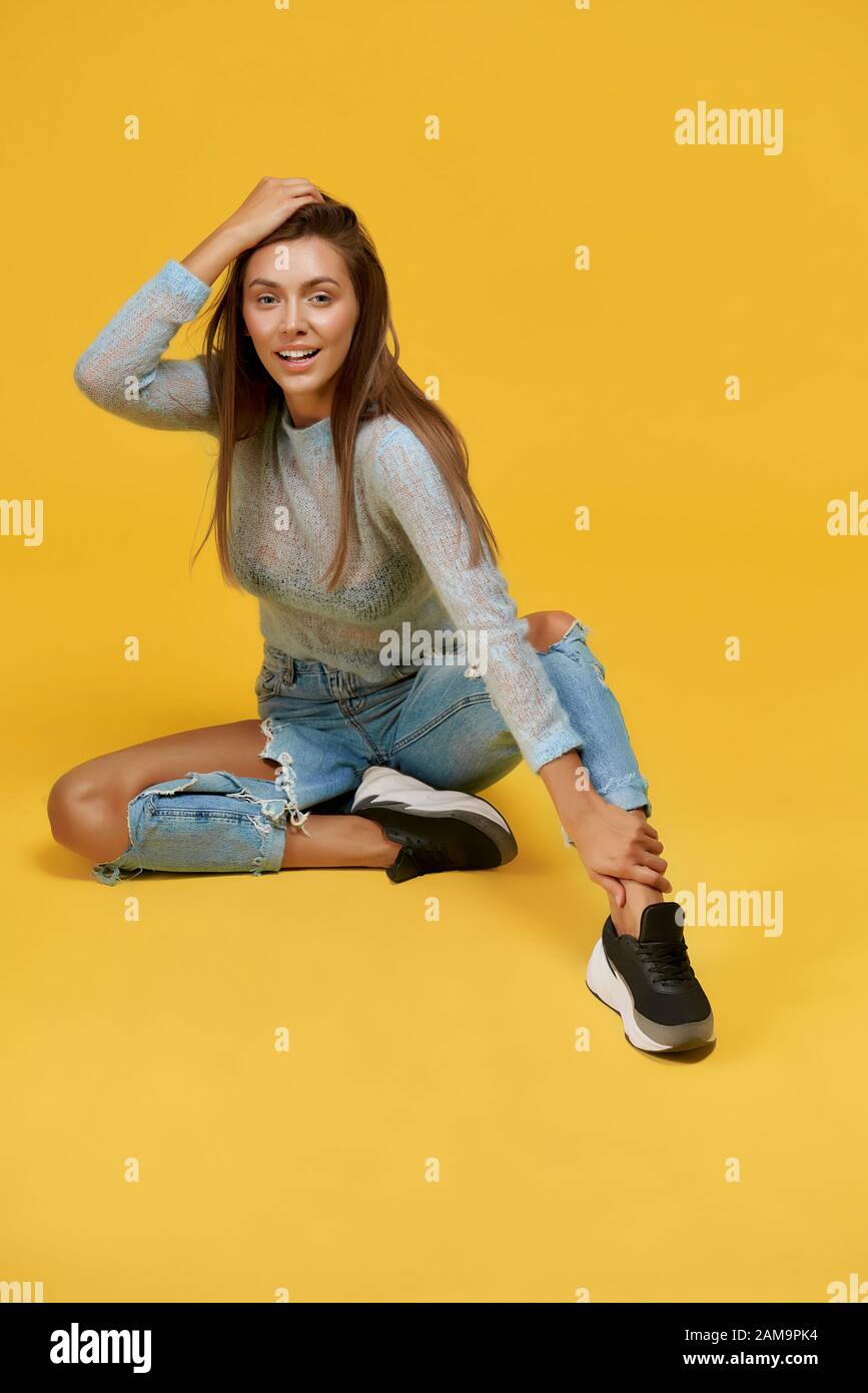 Vue avant de la jolie jeune fille assise avec la jambe gauche vers l'avant, touchant la cheville avec la main gauche, mettant la paume droite sur la tête, ouverture bouche portant le haut bleu avec manches longues, jeans déchiré. Banque D'Images