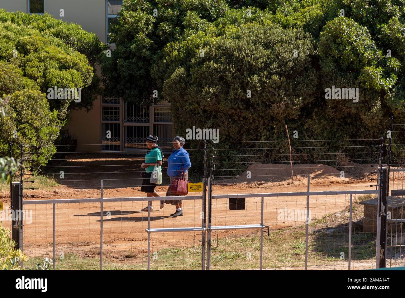 Le Cap Occidental, Afrique Du Sud, Décembre 2019. Deux femmes noires qui marchent pour travailler sur une route de terre ont une clôture de sécurité. Banque D'Images