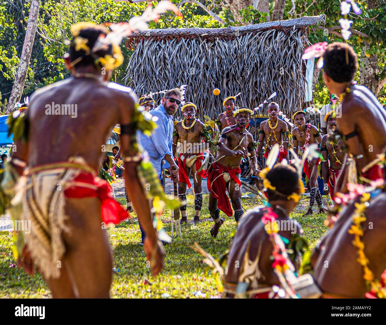 Un passager du navire d'expédition True North lance la balle orange lors d'une partie de cricket de style des îles Trobriand à Kwebwaga, îles Trobriand, Papouasie-Nouvelle-Guinée, un danseur costumé court vers lui, des hommes costumés se tiennent autour du guichet devant une hutte couverte de palmes. Banque D'Images