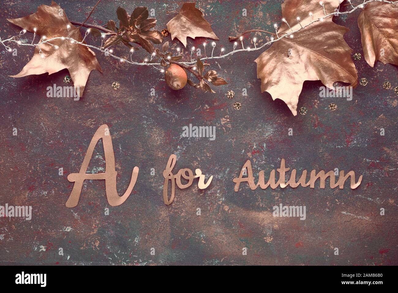 Plat, vue sur le dessus avec feuilles dorées peintes, guirlande lumineuse festive sur fond sombre texturé. Texte « A ou automne » en italique joint doré Banque D'Images