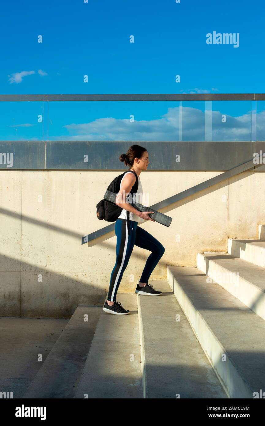 Femme sportive marchant sur des marches portant un tapis d'exercice et un sac à dos portant des vêtements de sport. Banque D'Images