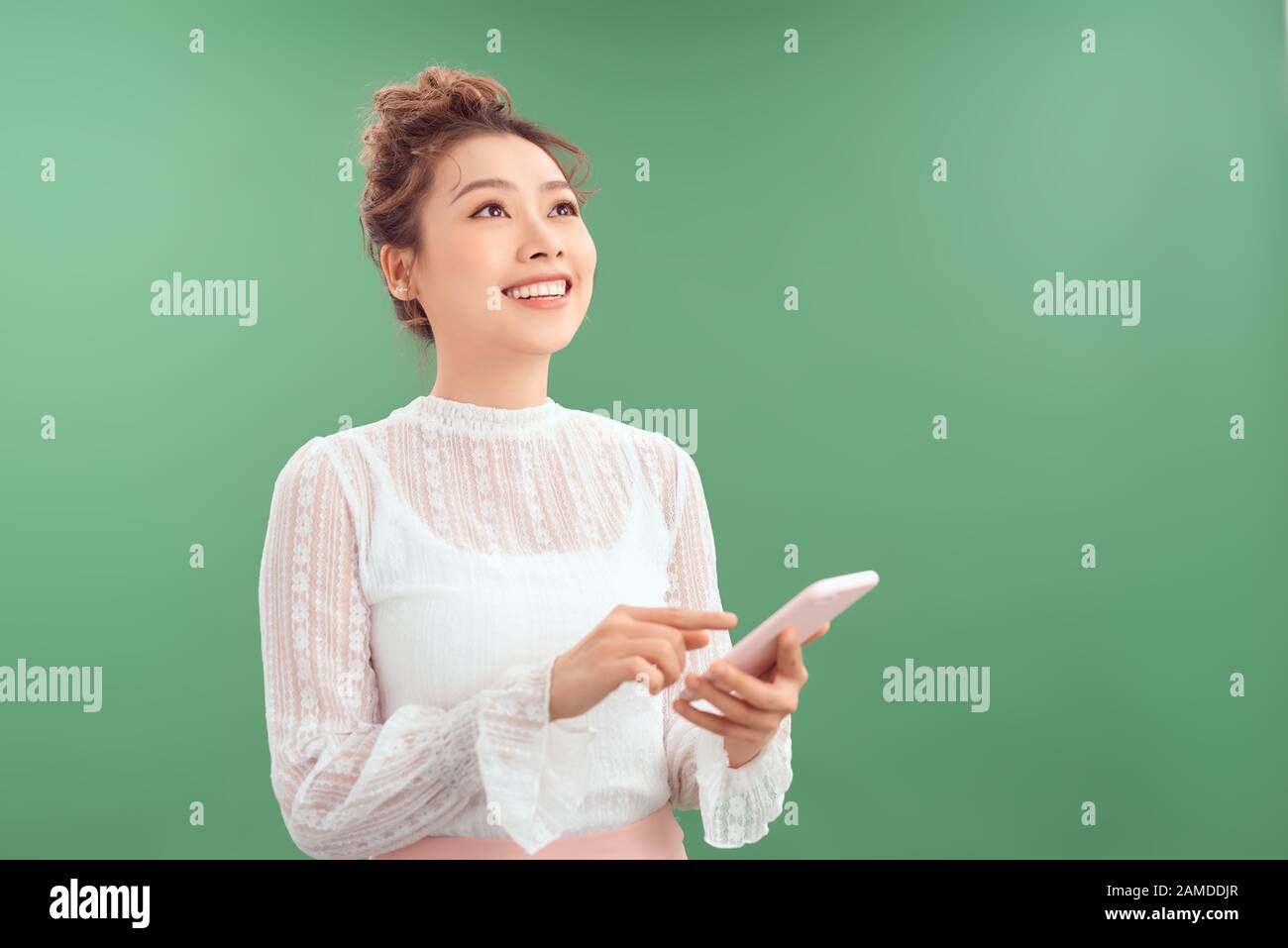 Portrait d'une jeune femme asiatique heureuse tenant un téléphone mobile tout en se tenant debout et en regardant isolé sur fond vert. Banque D'Images