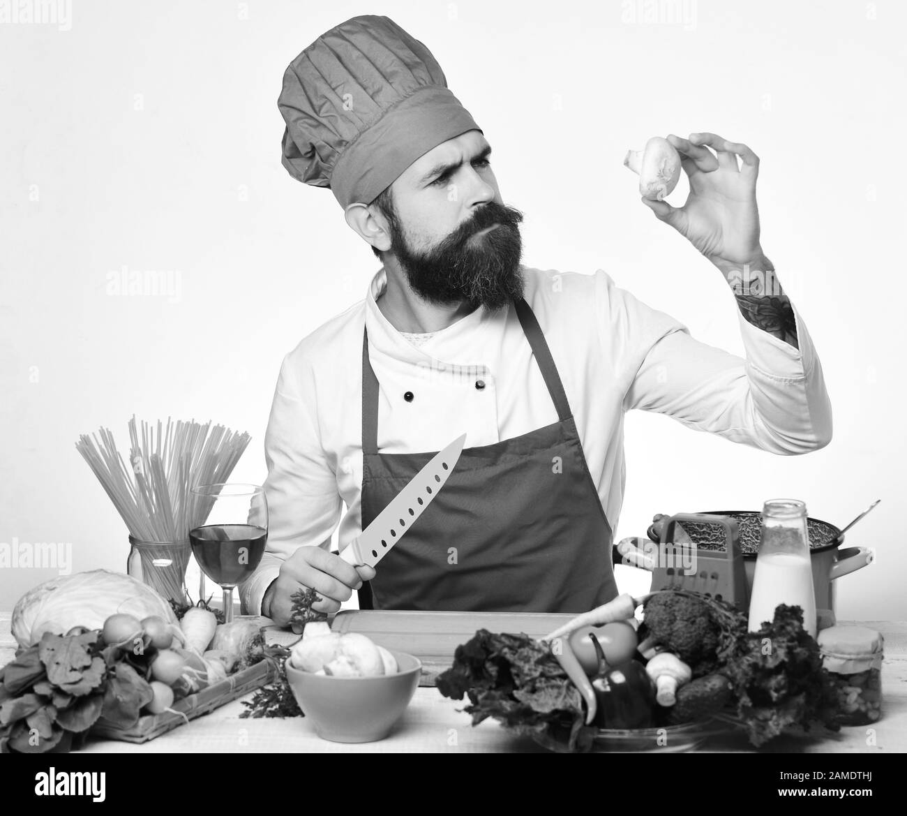Concept du processus de cuisson. Le chef prépare le repas. Homme avec barbe, tenez les champignons et le couteau sur fond blanc. Faites cuire avec le visage délicat en uniforme bordeaux assis par une table de cuisine avec des légumes et des ustensiles de cuisine. Banque D'Images