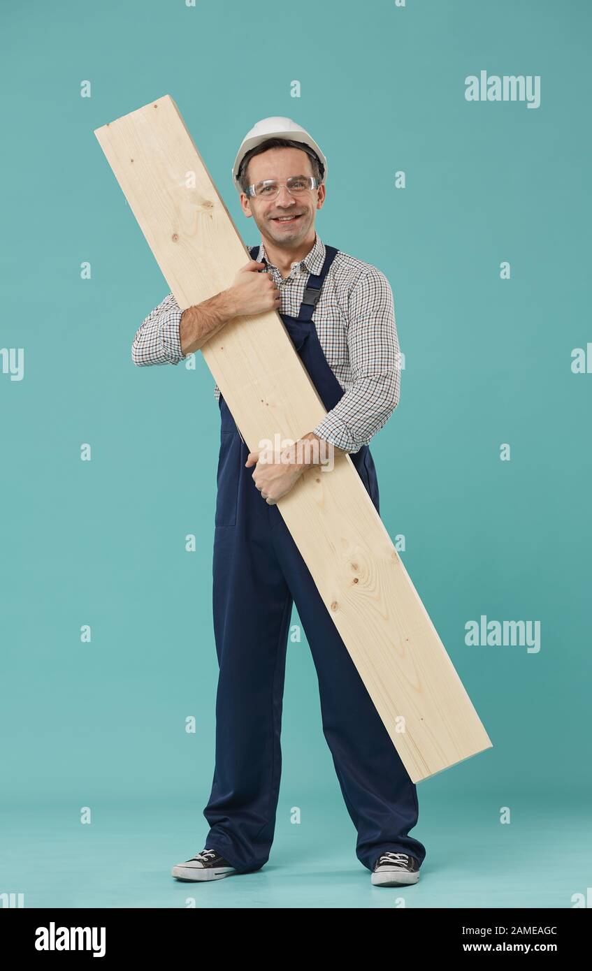 Portrait complet d'un travailleur professionnel ou d'un constructeur souriant à l'appareil photo tout en tenant du bois debout sur fond bleu Banque D'Images