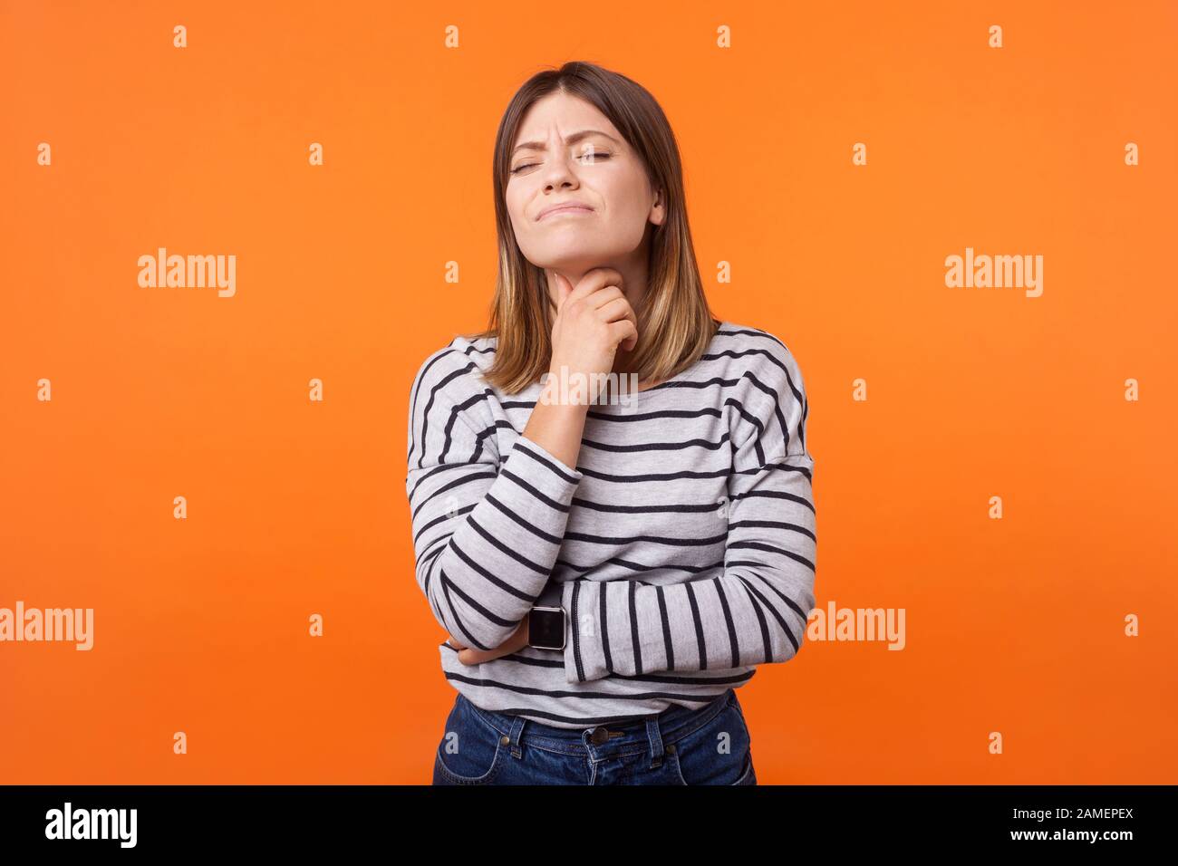 Portrait d'une femme malade avec des cheveux bruns dans une chemise rayée à manches longues se tenant au cou et froissant de la douleur, souffrant de mal de gorge, symptôme de grippe. Banque D'Images